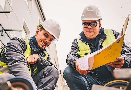 Bild mit zwei Personen kniend auf einer Baustelle in Arbeitskleidung mit Warnweste 