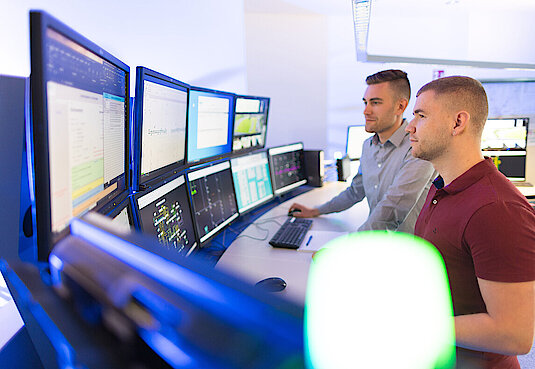 zwei männliche Mitarbeiter stehend an einem Schreibtisch vor mehreren Bildschirmen in der Leitstelle
