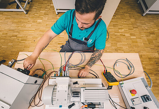 Bild mit einer Person in Arbeitskleidung sitzend an einem technischen Arbeitsplatz Elektrotechnik 